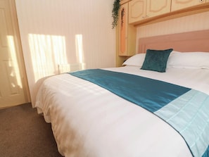 A neatly made bed with a white and teal bedspread, a wooden headboard, and a window with curtains. - 2br-garden-freeparking-charitydonation (Redruth)