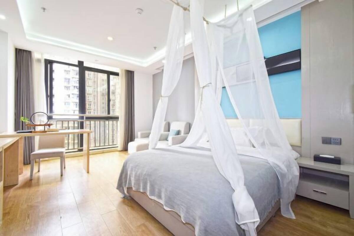 A bedroom with a bed, a desk, and a view of buildings through a large window.