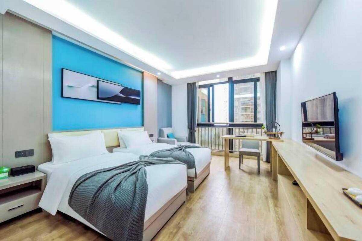 A modern hotel room with two beds, a desk, a chair, a TV, and a balcony with a city view.
