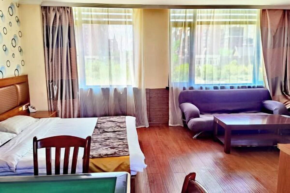 A hotel room with a bed, a wooden chair, a sofa, a coffee table, and large windows with curtains.