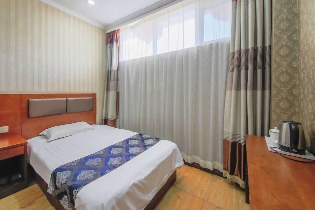 A hotel room with a bed, a wooden nightstand, a desk with a kettle, and a window with curtains.
