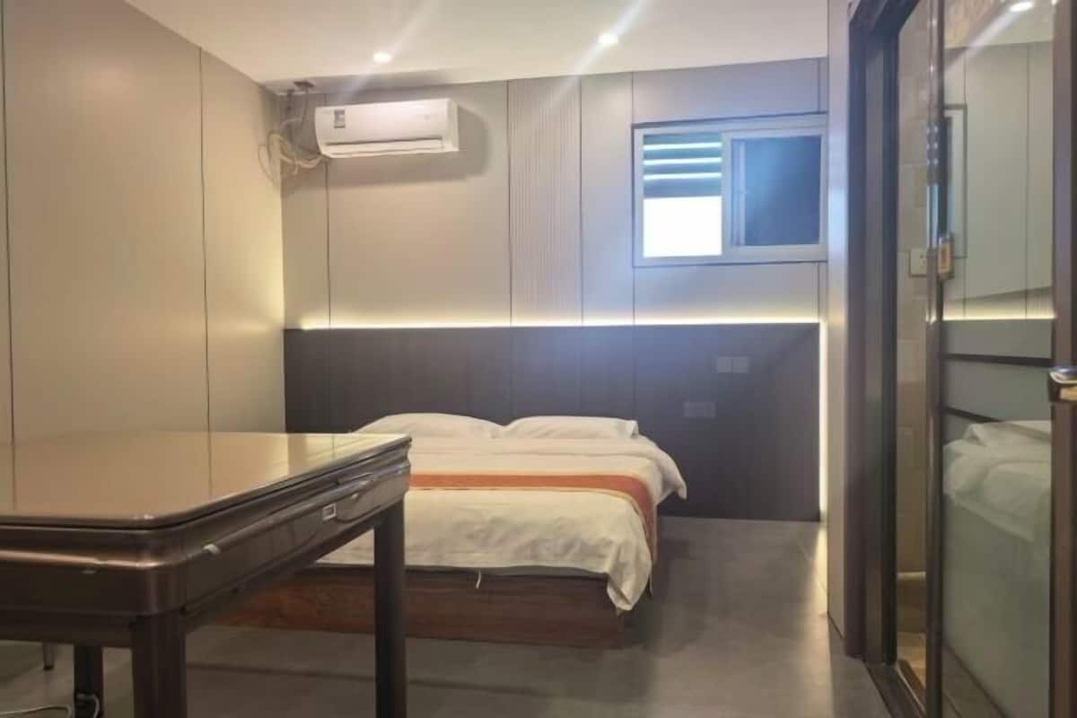 A hotel room with a bed, bedside table, air conditioning unit, and a window.