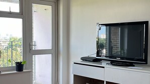 A modern living room with a flat-screen TV on a white console, wooden flooring, and a large window with a view of buildings. - Views from the 8th (London)