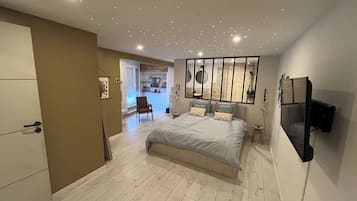 Een moderne slaapkamer met een groot bed, een aan de muur bevestigde televisie, en een badkamer zichtbaar achter een glazen scheidingswand.