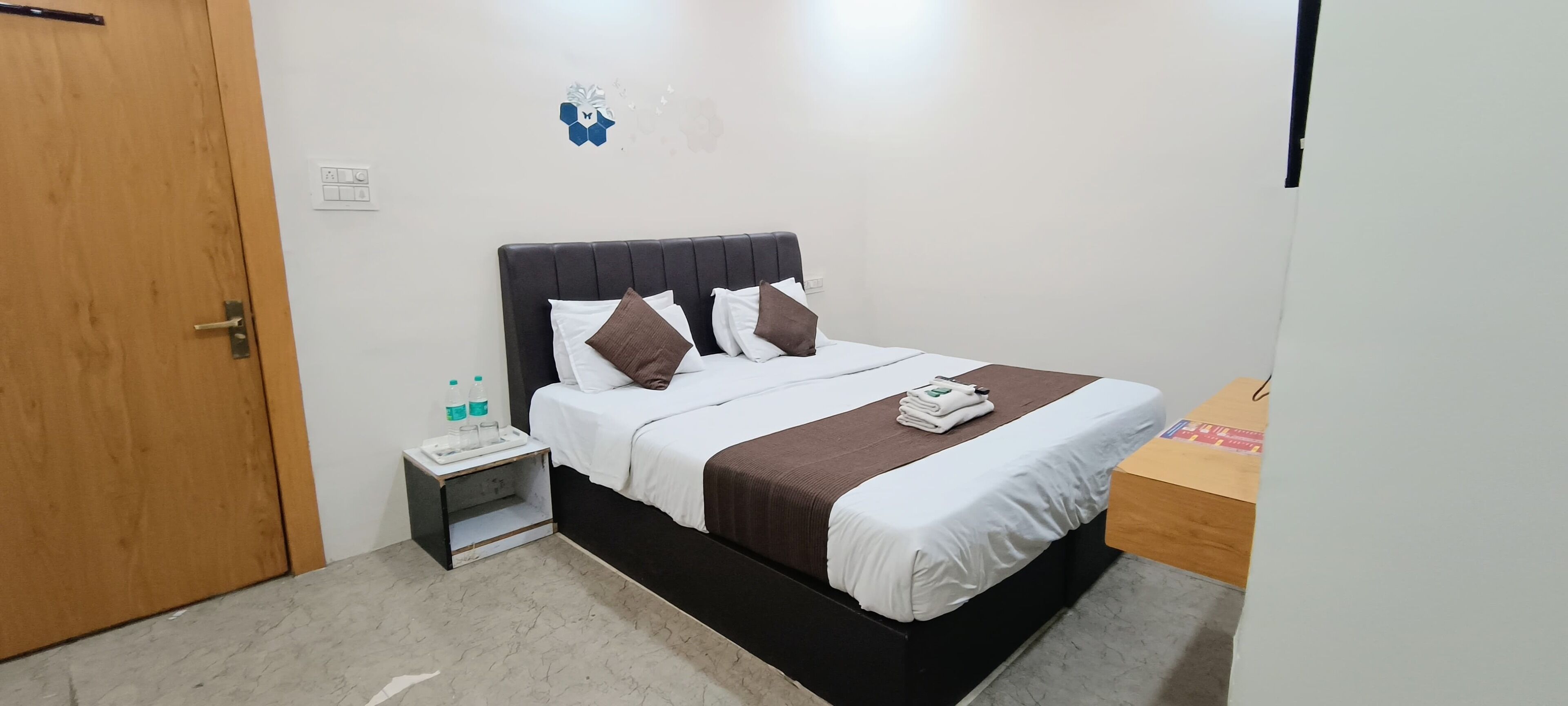 Una cama bien hecha con ropa de cama blanca y marrón, un cabecero de madera y una mesita de noche con una lámpara y una botella.