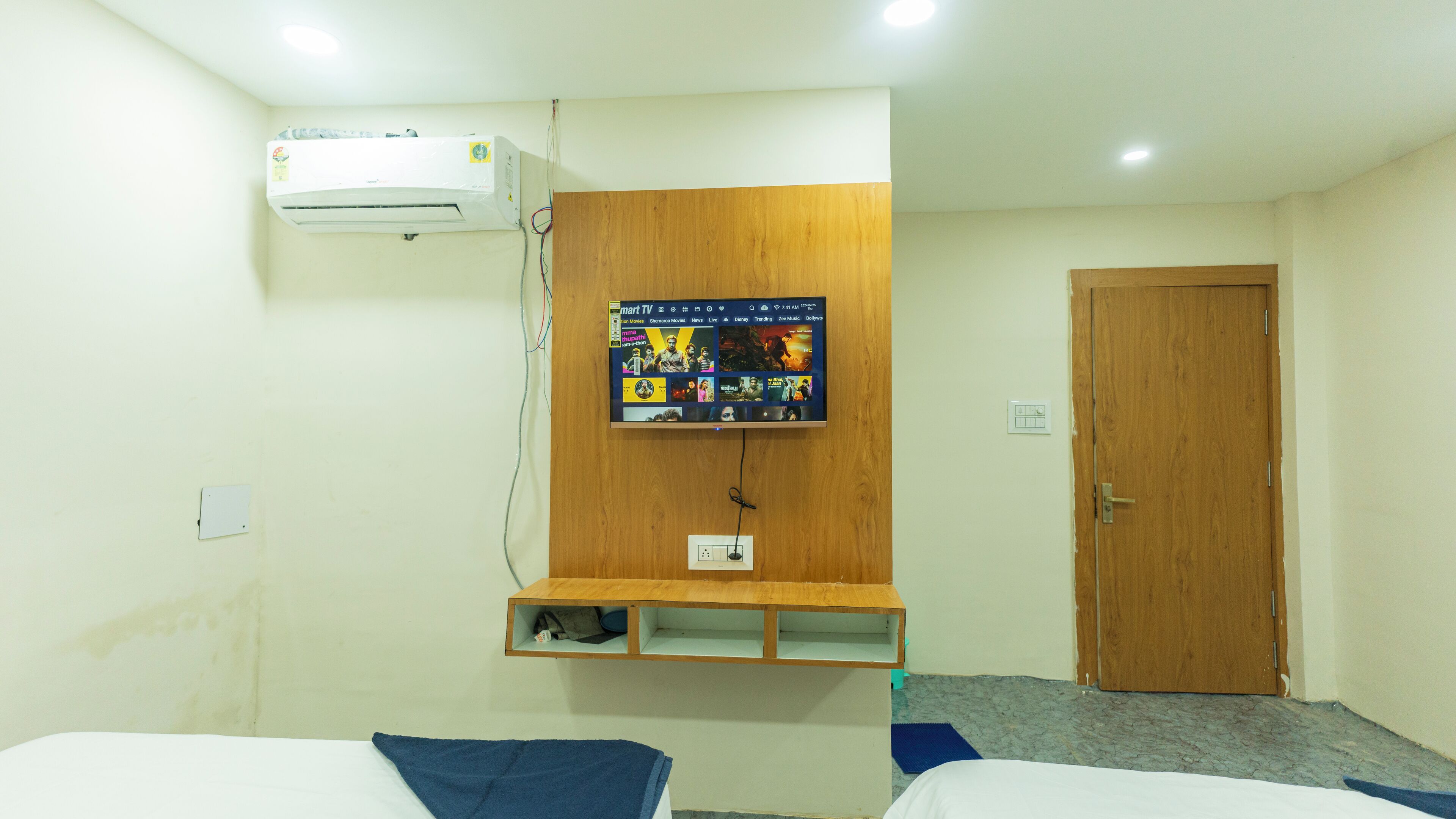 Ein Hotelzimmer mit einem an der Wand befestigten Fernseher, einem Holzregal, einer Tür und einem Bett.