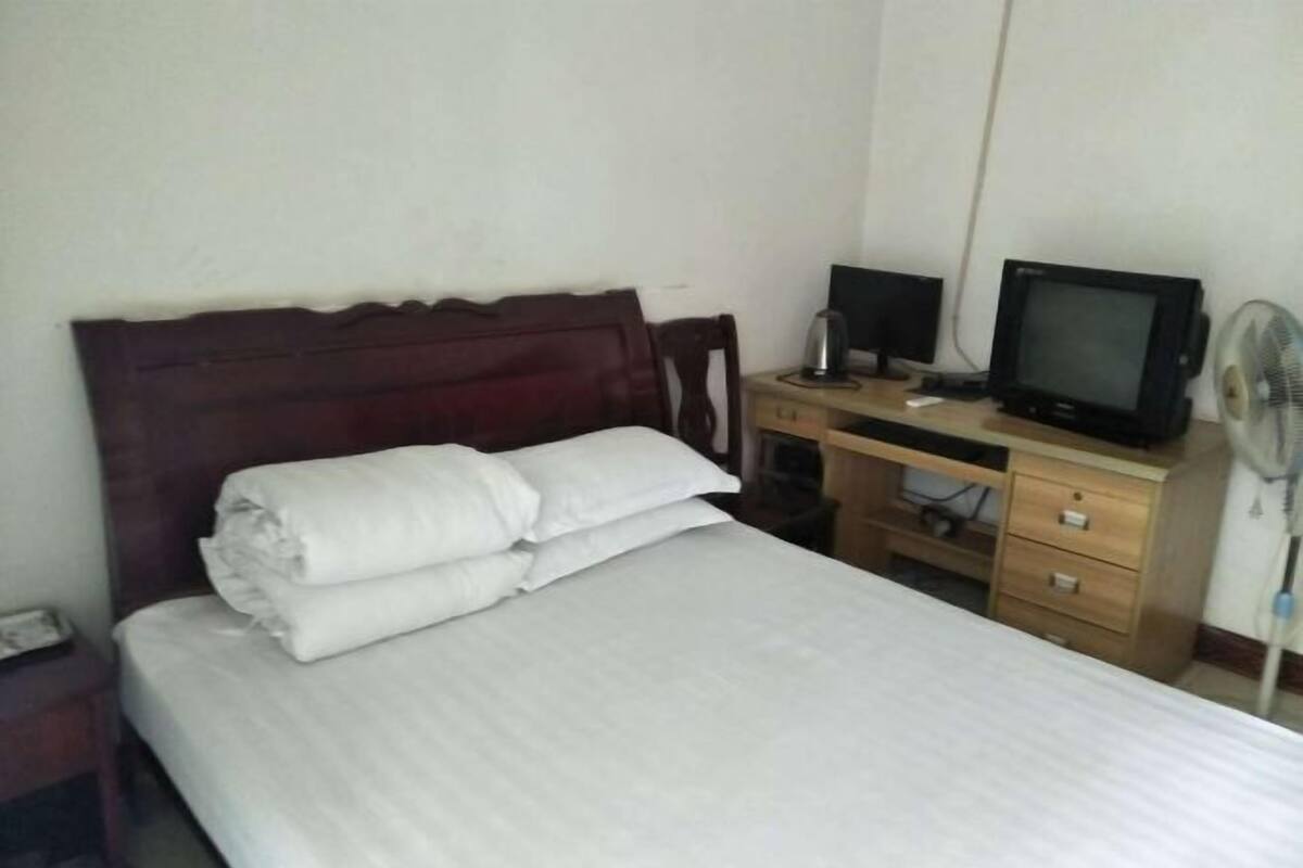 Une chambre d’hôtel avec un lit, un bureau en bois équipé d’une télévision et d’un ventilateur, et une petite table de chevet.