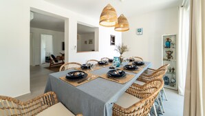 Un coin repas avec une longue table dressée pour six personnes, entourée de chaises en osier, sous trois lampes en osier suspendues au plafond.