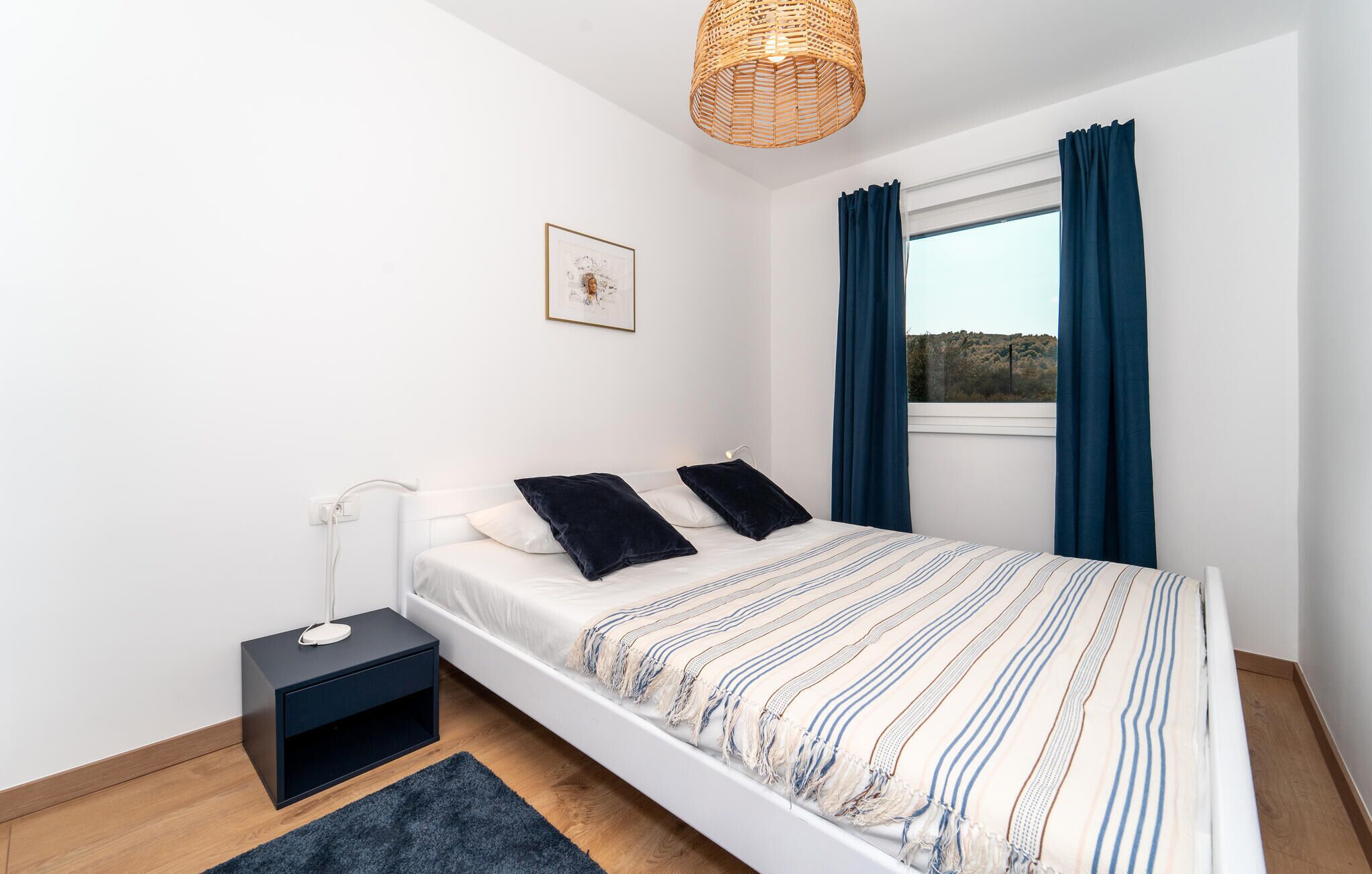 Een slaapkamer met een bed, nachtkastje, lamp, vloerkleed en een raam met gordijnen.