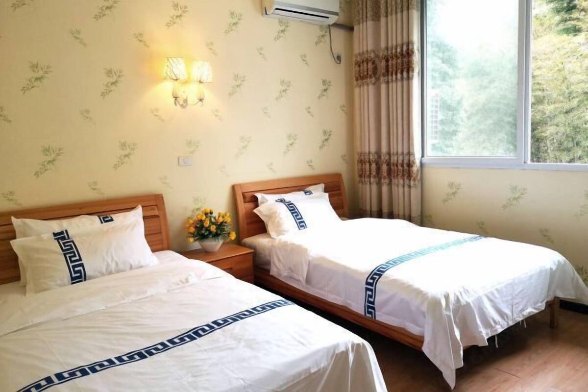 Habitación de hotel con dos camas, una mesita de noche con un florero y aire acondicionado de pared, además de una ventana con cortinas.
