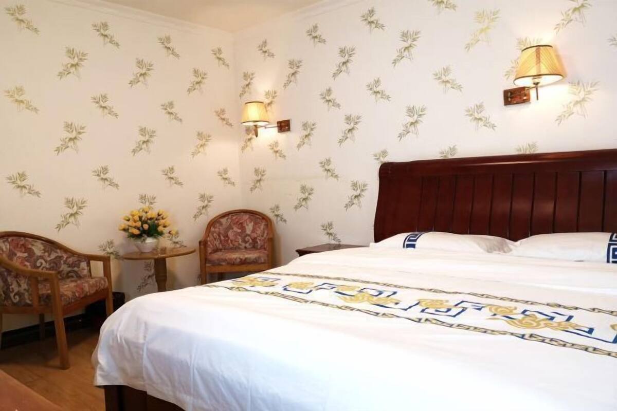 Habitación de hotel con una cama grande, dos sillones, una mesita con un florero y lámparas fijadas a la pared.