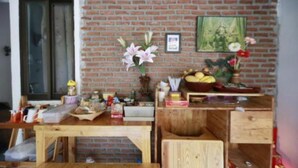 A room with a brick wall, a wooden desk, and various items on the desk including a vase with flowers, a framed picture, and a basket of fruit. - Banana Hostel Donmuang (Bangkok)