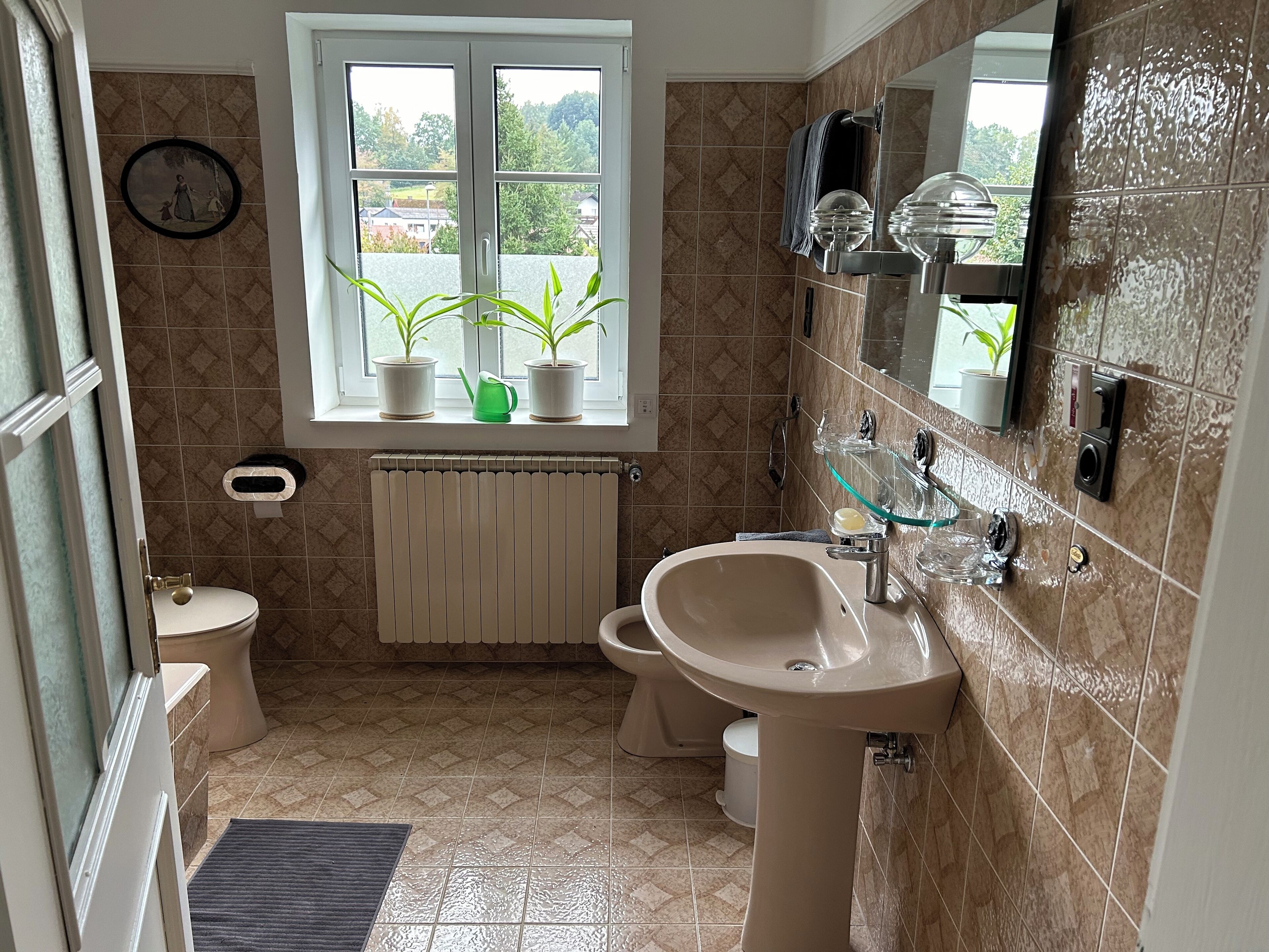 Une salle de bain équipée de toilettes, d’un bidet, d’un lavabo et d’une fenêtre agrémentée de plantes en pot.