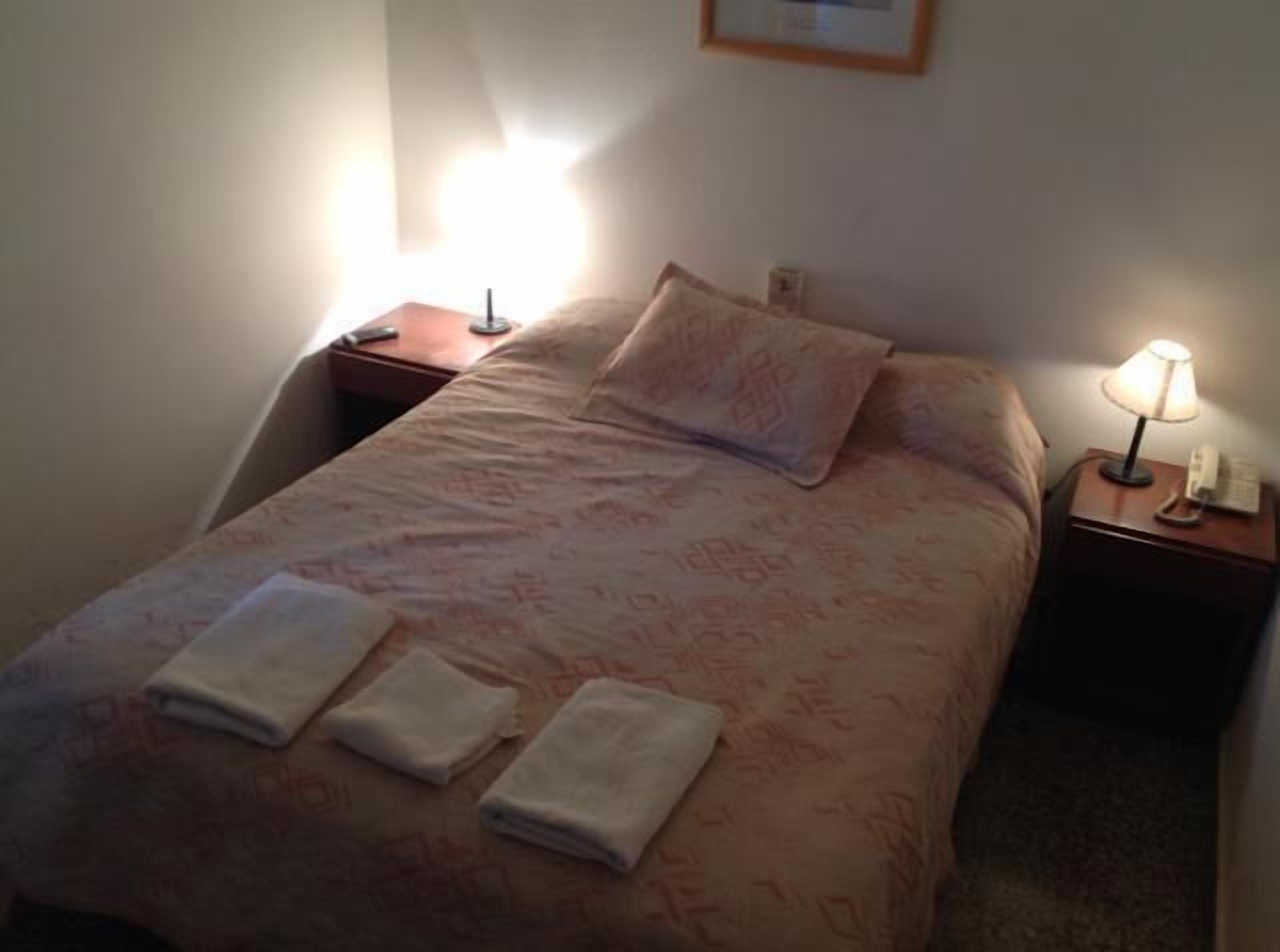 Ein ordentlich bezogenes Bett mit einem gemusterten Überwurf, zwei Kissen und zwei gefalteten Handtüchern. Rechts vom Bett befindet sich eine Nachttischlampe, links ein kleiner Tisch mit Lampe und Telefon.