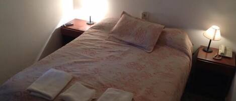 Ein ordentlich bezogenes Bett mit einem gemusterten Überwurf, zwei Kissen und zwei gefalteten Handtüchern. Rechts vom Bett befindet sich eine Nachttischlampe, links ein kleiner Tisch mit Lampe und Telefon.