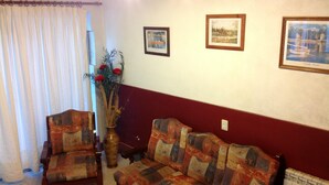 Una sala de estar con un sofá, un sillón y un jarrón con flores. Hay tres cuadros en la pared.