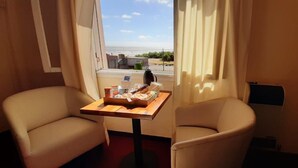 Ein Hotelzimmer mit zwei beigen Sesseln, einem kleinen Holztisch mit Frühstücksangeboten und einem Fenster mit Blick auf Strand und Gebäude.