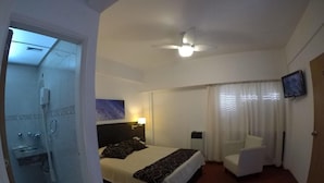 Ein Hotelzimmer mit Bett, Sessel, Fernseher, Deckenventilator und Duschbereich.
