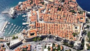 Vista aérea de uma vila costeira com uma muralha fortificada, um porto com barcos e um conjunto denso de edifícios com telhados vermelhos.