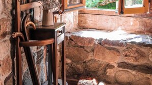 A rustic stone interior with a wooden chair, a small table with a vase, and a window with a view of greenery.