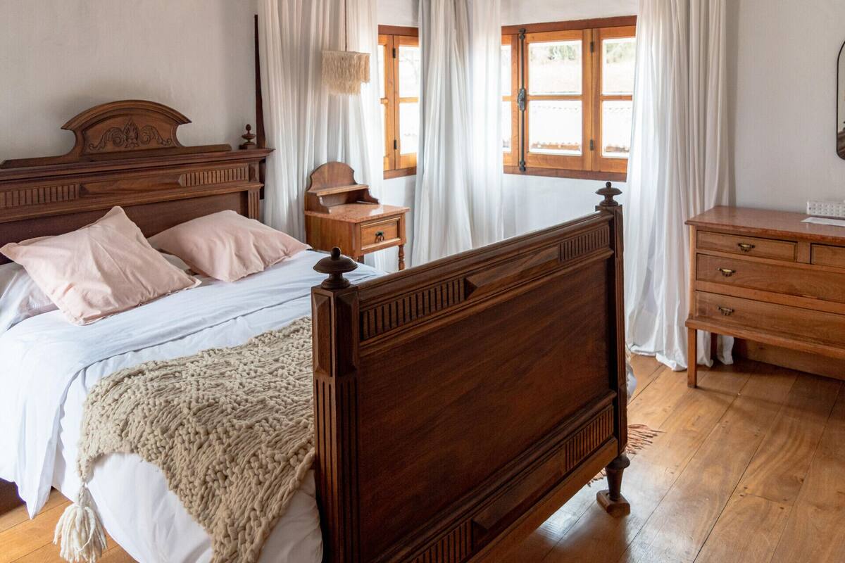 A bedroom with a wooden bed, a bedside table, a dresser, and a window with curtains.
