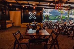 A restaurant with wooden tables and chairs, a stage with a neon 'X' sign, and outdoor seating with umbrellas. - Blu Terrace Hotel (Blumenau)