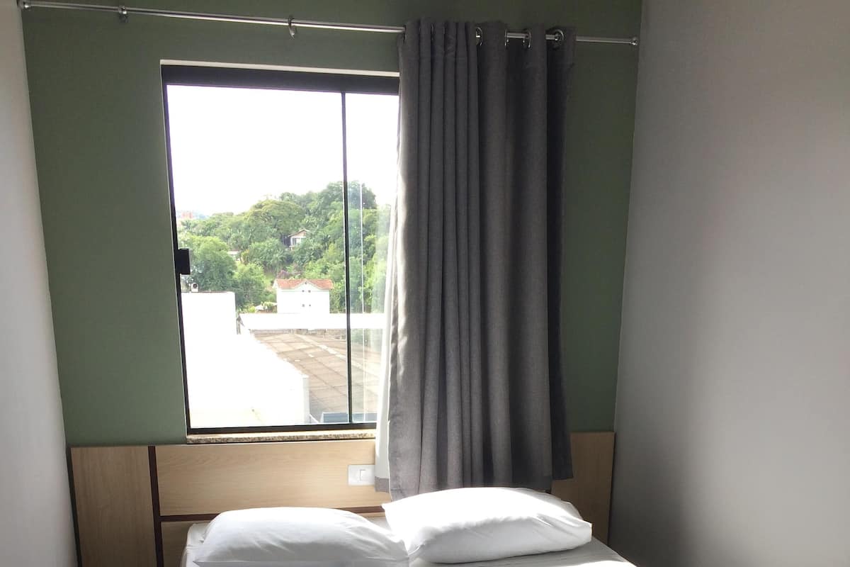 A single bed with white linens, a plastic bag on the bed, and a window with a curtain.