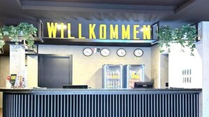 A reception desk with a 'WILLKOMMEN' sign, clocks, and a refrigerator. - Blu Terrace Hotel (Blumenau)