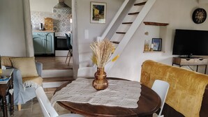 Un comedor con una mesa redonda de madera, sillas blancas y un jarrón con flores secas. Una escalera conduce a una cocina con baldosas estampadas.