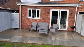 A brick house with a tiled patio, two wicker chairs, a barbecue, and a sliding glass door. - Newly Renovated 3 bed House Near Beach in Heacham (King's Lynn)