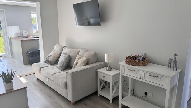A modern living room with a grey sofa, a white side table, a TV mounted on the wall, and a small white desk with drawers. - Newly Renovated 3 bed House Near Beach in Heacham (King's Lynn)