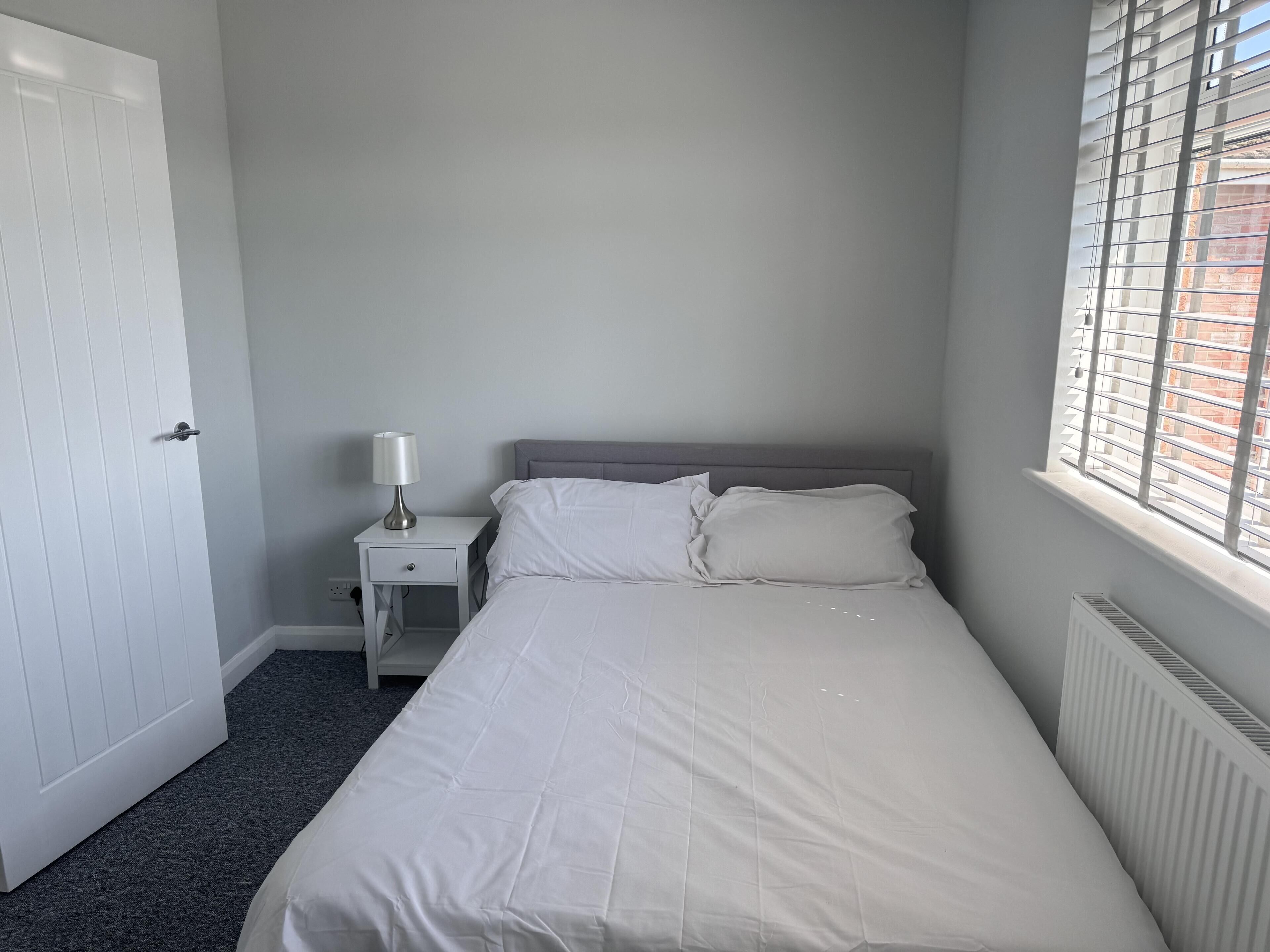 A bedroom with a bed, a nightstand, a lamp, a window with blinds, and a radiator.