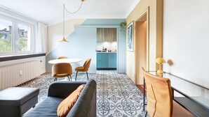 Un salon moderne comprenant un canapé, une table à manger avec des chaises, un coin cuisine et une grande fenêtre laissant entrer la lumière naturelle.