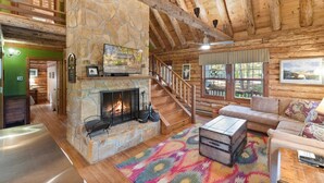 A log cabin interior with a stone fireplace, wooden staircase, and a cozy living area with a sofa and a colorful rug. - Log Cabin w/ Hot Tub & Waterfront Patio! (Harpers Ferry)
