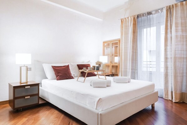 A hotel room with a bed, bedside tables, a nightstand, a lamp, a tray with towels, and a window with curtains.