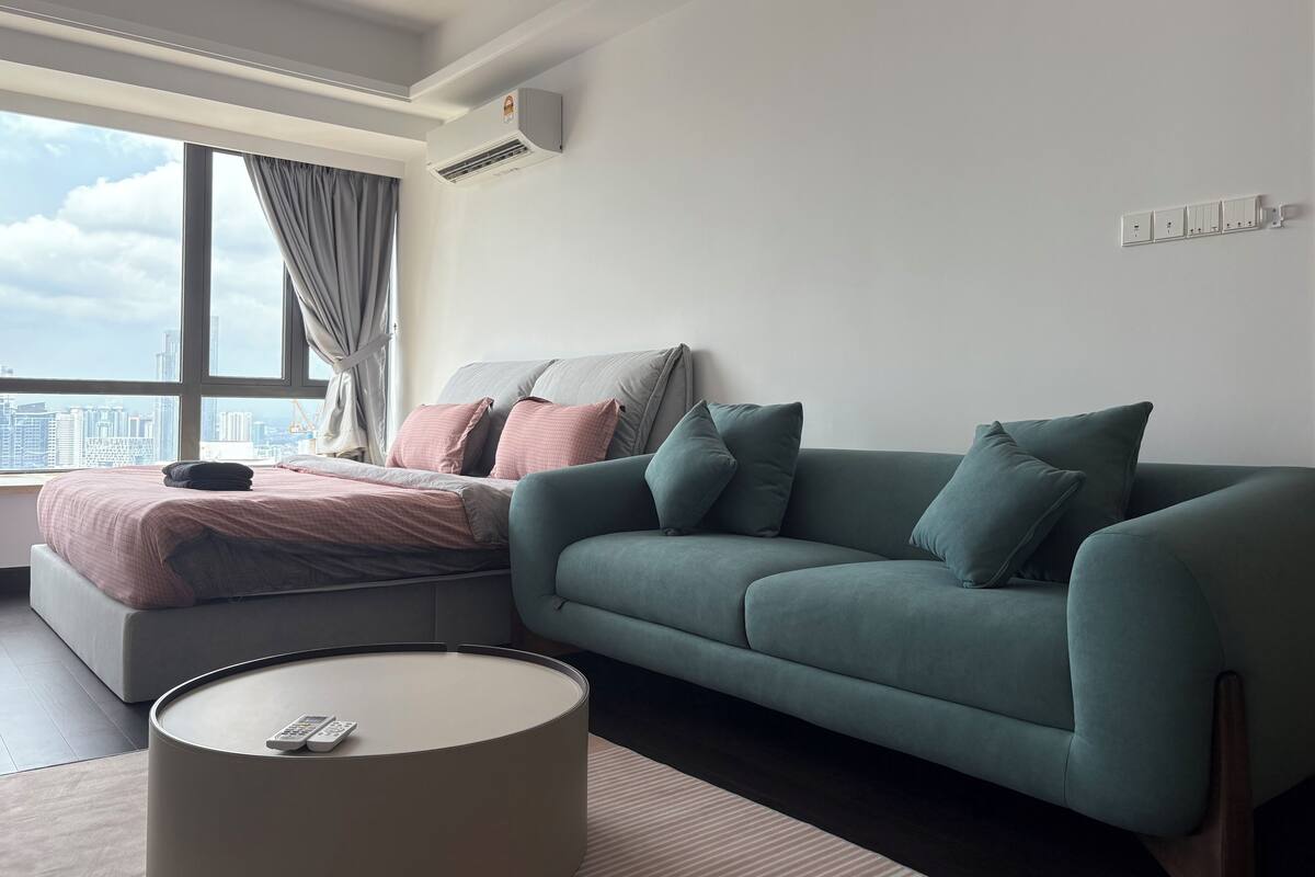 A modern bedroom with a bed, a green sofa, a round coffee table, and a large window with curtains.