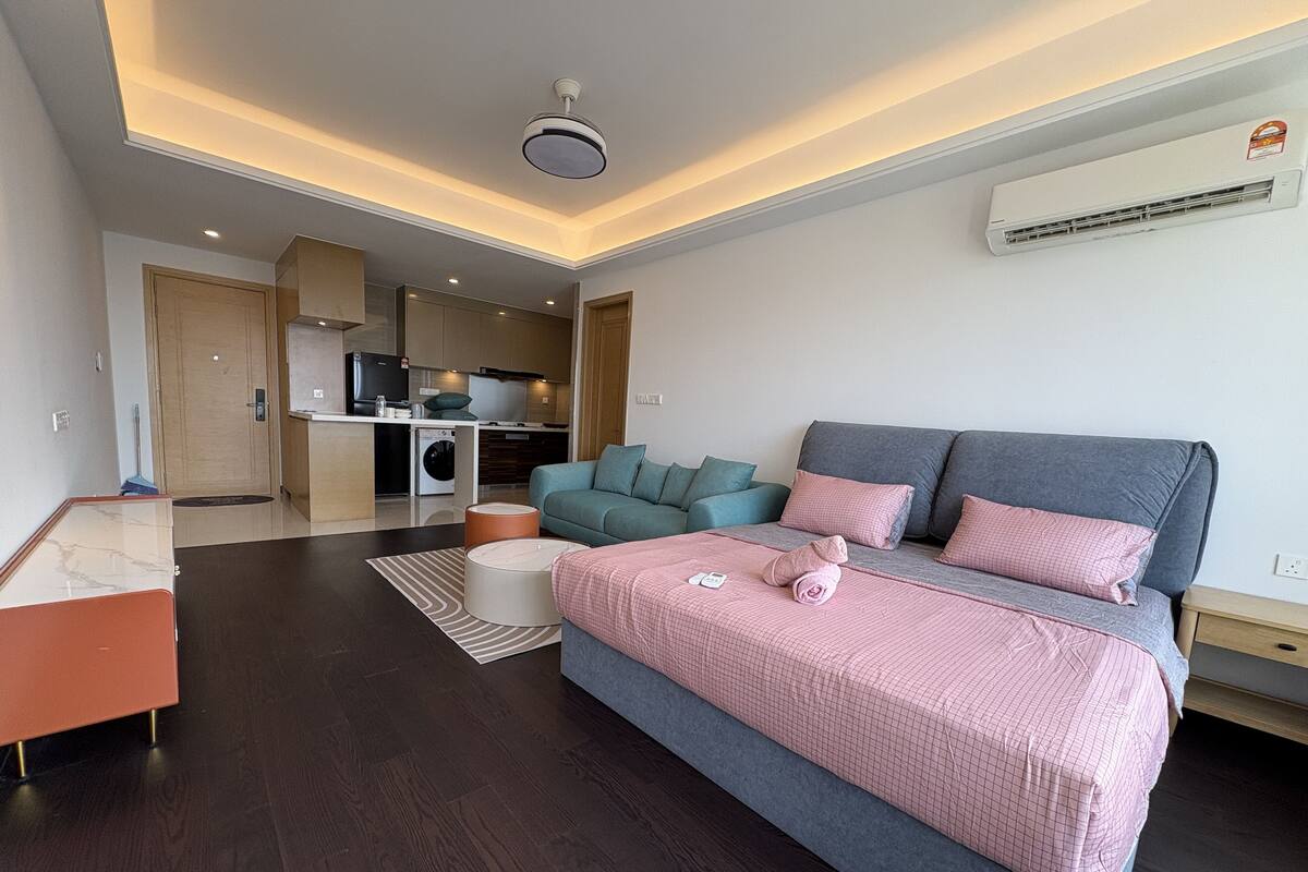 A modern hotel room with a large bed, a sofa, a small table, and a kitchenette in the background.