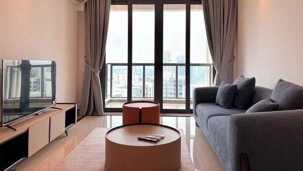 A modern living room with a large window, a flat-screen TV, a grey sofa, and a round coffee table. - R&F Seine Region (Johor Bahru)