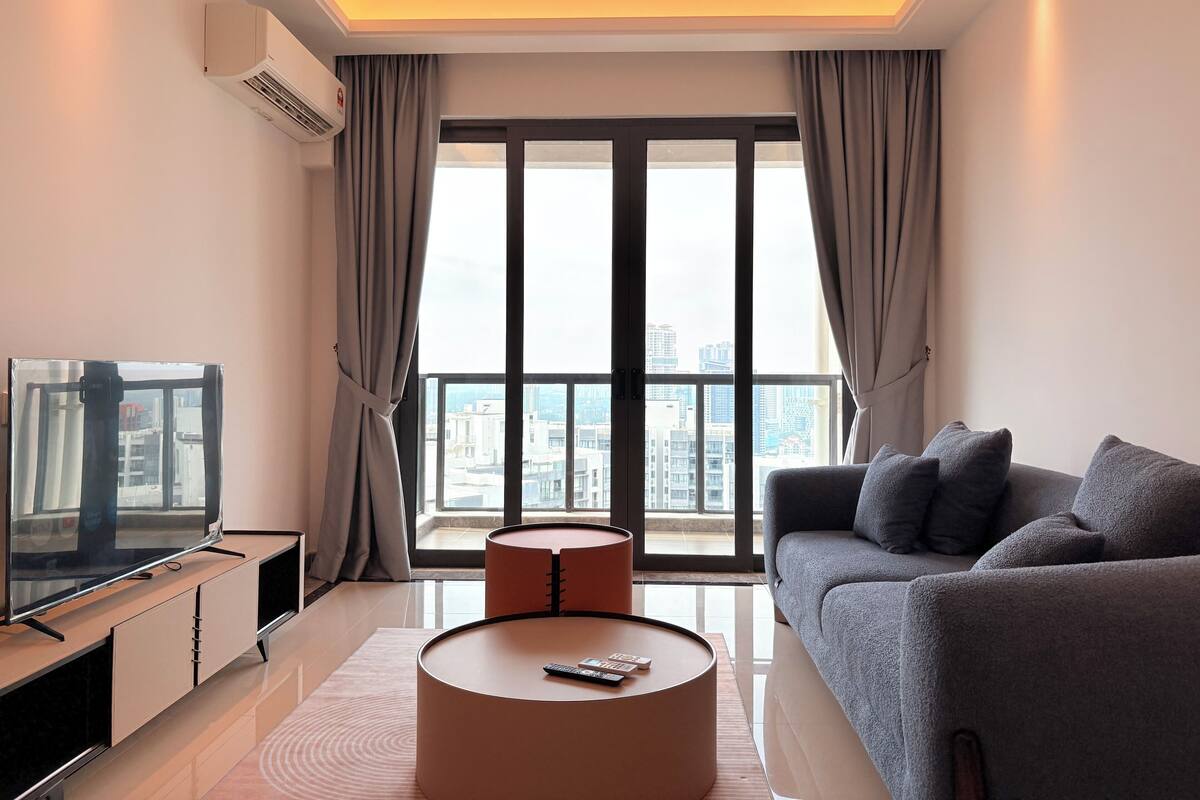 A modern living room with a large window, a flat-screen TV, a grey sofa, and a round coffee table.