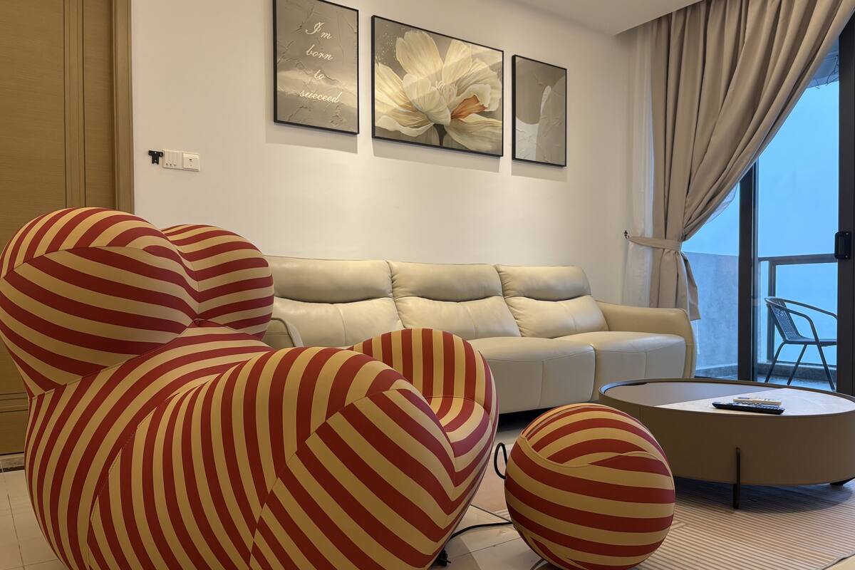 A modern living room with a beige sofa, striped armchairs, and a round coffee table. There are three framed artworks on the wall and a view of the outdoors through large windows.