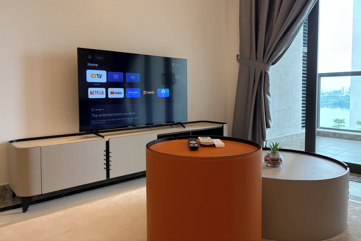 A modern living room with a large flat-screen TV mounted on a wall, a small round table with a remote control, and a view of a body of water through a large window.