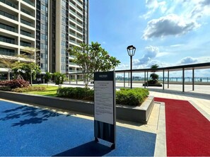 Un espace extérieur aux couleurs bleu et rouge, agrémenté d’un panneau indicateur, entouré de verdure et avec un immeuble de grande hauteur en arrière-plan.