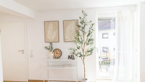 Una habitación luminosa con una puerta blanca, una mesa consola de vidrio transparente, dos obras de arte con motivos de hojas, una planta en maceta y vistas a un balcón exterior a través de un ventanal amplio.