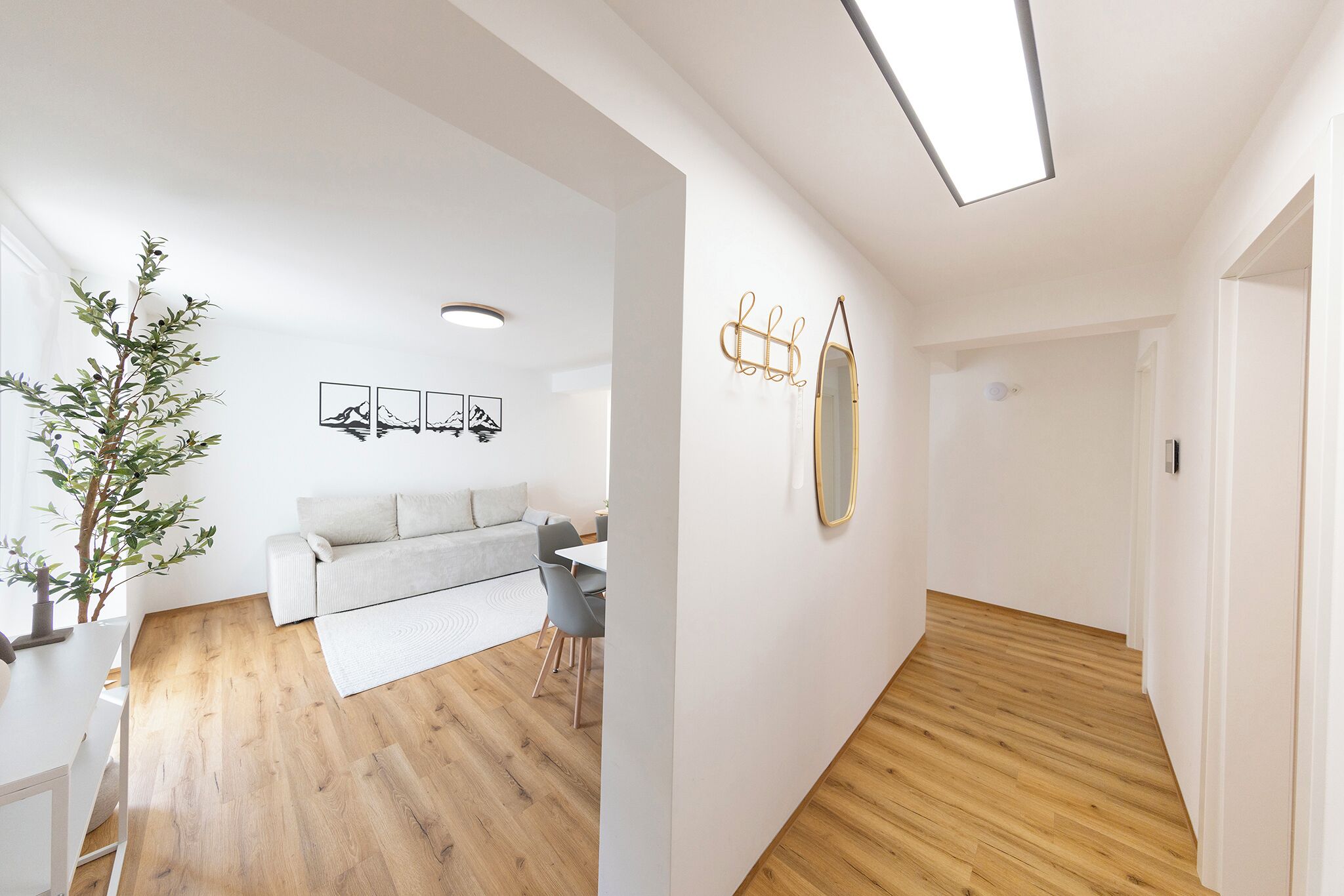 Un salon moderne avec un canapé, une table à manger et des chaises. On y trouve une plante, une œuvre d’art accrochée au mur et un miroir. Le sol est en bois et les murs sont peints en blanc.