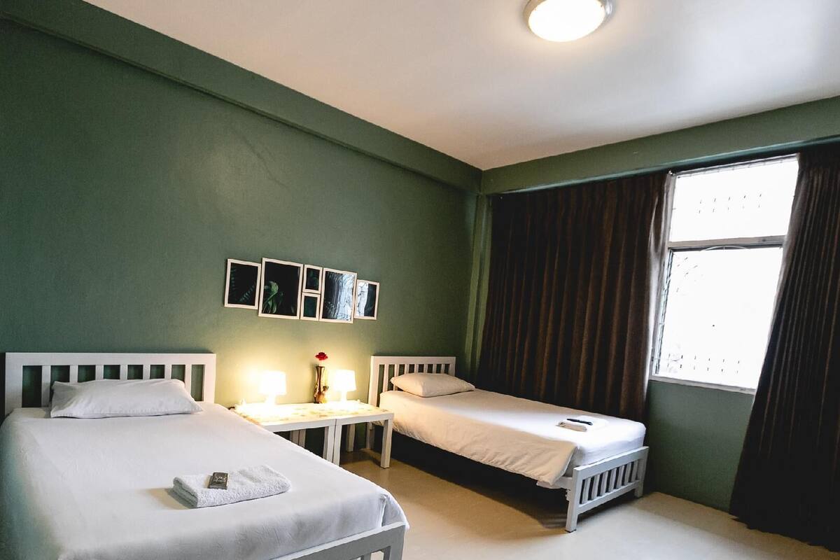 A hotel room with two single beds, a small table with a lamp, and a window with curtains.