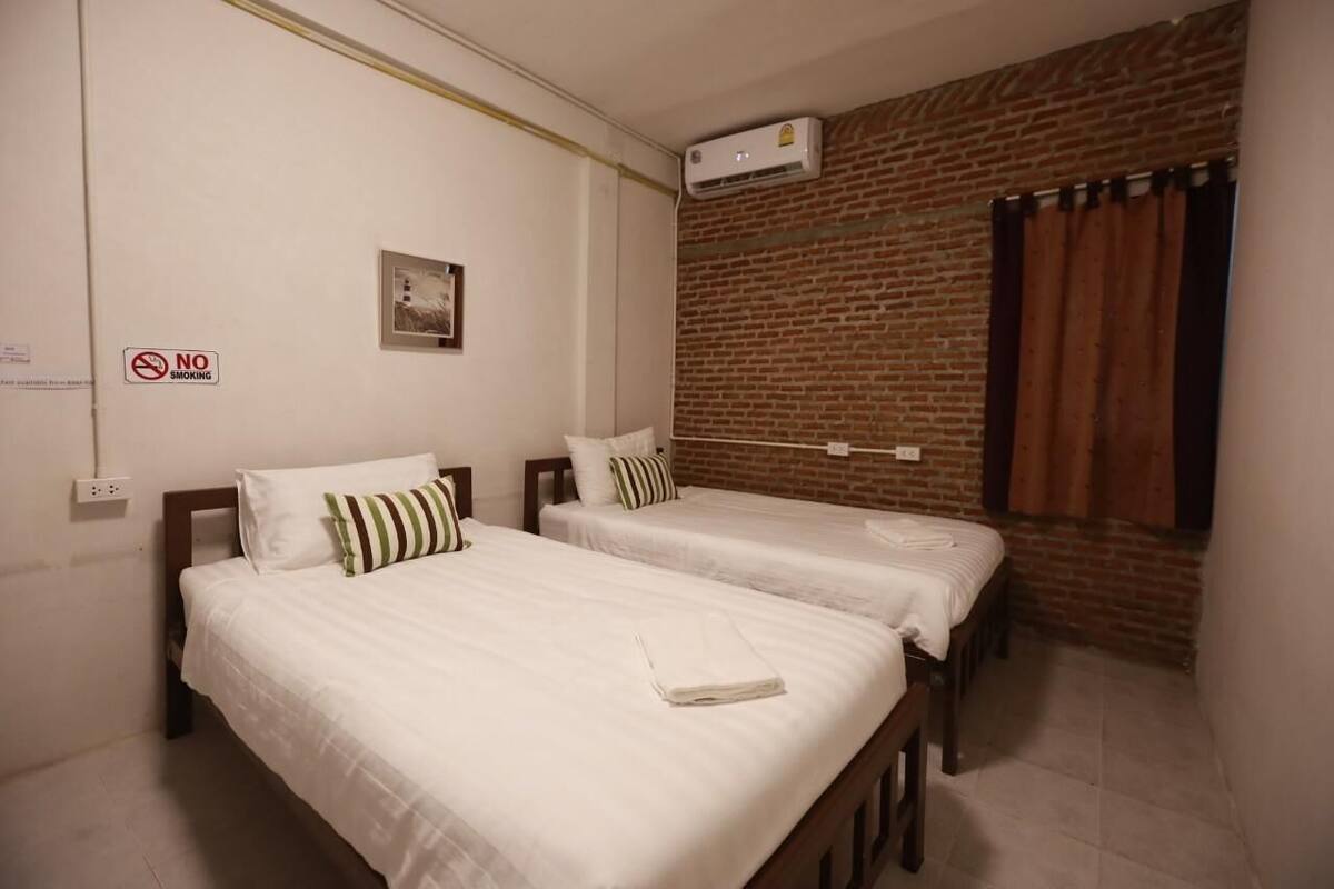 A hotel room with two single beds, a brick wall, an air conditioning unit, and a 'No Smoking' sign.