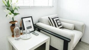 A minimalist living room with a white sofa, a small white table, and a window with blinds. - Sleepcase Hostel (Bangkok)