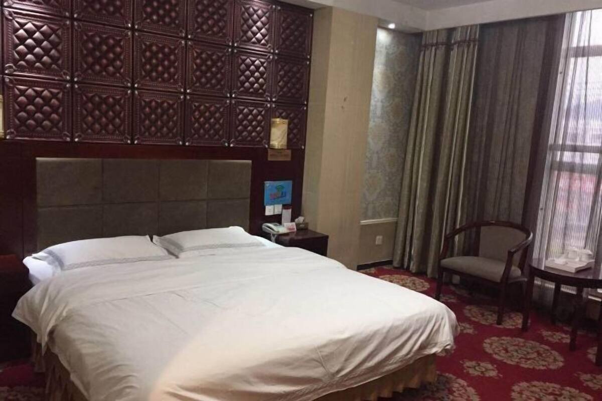 Habitación de hotel con una cama grande, un cabecero de madera, una silla, una mesita y una ventana con cortinas.