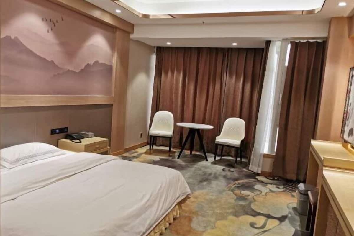 Habitación de hotel con cama, dos sillas, una mesita, un cuadro en la pared y ventana con cortinas.