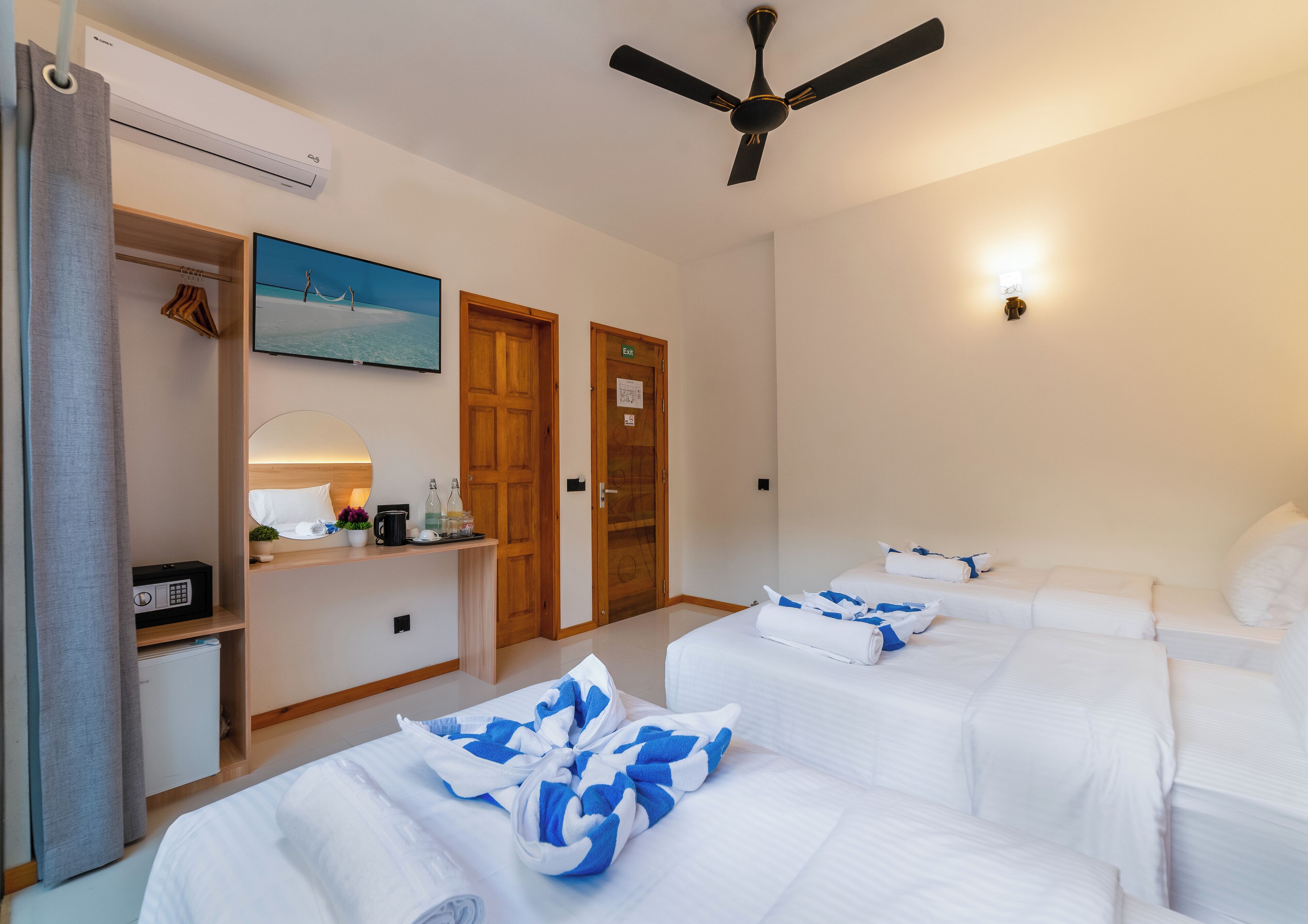 Habitación de hotel con dos camas, un escritorio con espejo, un ventilador de techo y un cuadro en la pared.